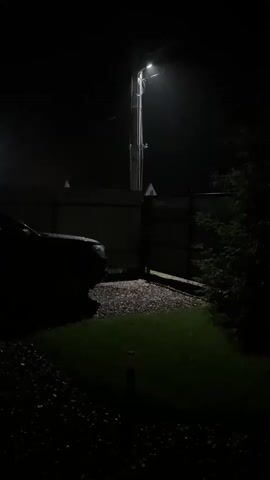 A brief video clip shows a dark outdoor scene. A single street light illuminates a section of a gravel driveway and a patch of grass. A dark-colored vehicle is partially visible on the left side of the frame. A dark fence runs horizontally across the middle ground. A portion of a tree is visible on the right side of the frame. The background is mostly black, suggesting it is nighttime or very dark.