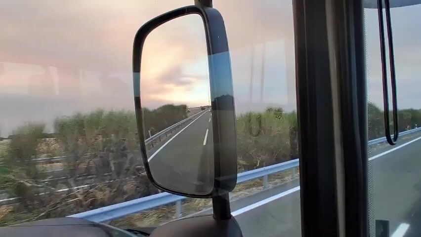 The video shows a view from inside a vehicle, likely a truck due to the presence of a large side mirror in the foreground. The side mirror reflects a road ahead, with a highway stretching into the distance. On either side of the road are green bushes and vegetation, with a metal guardrail running along the edge. The sky is filled with a soft, hazy sunset in shades of pink and orange. The sun is visible as a bright, diffused disc low in the sky. In the distance, beyond the vegetation, are fields of dry grass or crops, with scattered trees. The overall impression is of a scenic drive through a rural landscape at dusk. The vehicle appears to be moving, indicated by the slight blur of the surrounding environment. There are no people visible in the video.