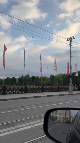 The video, filmed from the passenger seat of a vehicle, shows a view of a street with tram tracks. A decorative bridge with flags is visible to the left. The flags are alternately red and white. A cyclist is briefly seen on the bridge. A street lamp is standing on the sidewalk. The rearview mirror shows a building in the distance. The sky is cloudy with patches of blue.