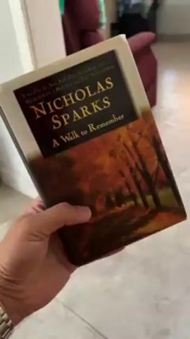 A hand holds the book "A Walk to Remember" by Nicholas Sparks. The book has a white cover and a photo of a tree-lined path with autumn colors. The hand places the book on a wooden surface. The hand opens the book to the title page.