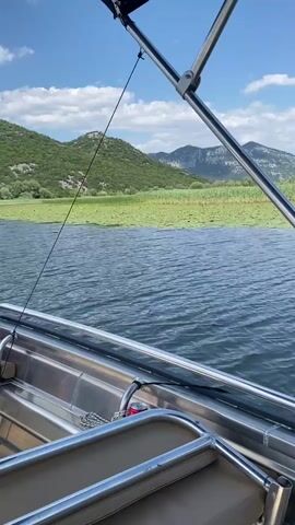 A first-person perspective video shows a boat moving through water. The camera is positioned inside the boat, looking out over the bow. The water is choppy with moderate waves. In the distance, there is a large area of green vegetation, possibly lily pads or reeds, on the water. Beyond the vegetation are rolling green hills covered in trees, with some bare rock faces visible. The sky is blue with scattered white clouds. The boat has a canopy supported by metal poles, and the edge of the boat with metal railings is visible in the foreground. The overall impression is a serene boat ride through a natural landscape.