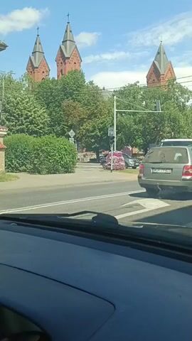 The video captures a street scene with a prominent church in the background, featuring three distinct spires. Trees and lush green bushes line the street, suggesting a pleasant, possibly summer, day with clear blue skies and scattered clouds. Two vehicles are visible in the foreground: a silver station wagon and a white flatbed truck. The station wagon is facing away from the camera, while the truck is positioned in front of it, also moving away. The truck appears to be carrying a load of pink flowers.

The perspective is from inside a car, looking forward through the windshield. The dashboard of the car is partially visible, framing the view. The scene unfolds at an intersection, indicated by the presence of a traffic light. The overall atmosphere seems peaceful and ordinary, with no obvious signs of distress.

The visible text on the license plate of the silver car is "MKZ 7905". Additionally, there is a brief Russian phrase spoken in the audio that translates to "Ukraine, rockets are not flying". This statement may be intended to express relief or a contrast between the current peaceful scene and potential conflict elsewhere. The context of Ostrów Mazowiecka, Poland, places the scene in a European setting. The time of day appears to be daytime, likely late morning or early afternoon, given the bright sunlight and clear sky.