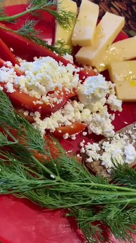 A close-up shot shows a red plate with food. The food includes slices of yellow cheese drizzled with oil, slices of red bell pepper topped with crumbled white cheese, a piece of dark rye crispbread spread with white cheese, and a garnish of fresh green dill. The background is blurred but appears to be a surface with a coffee bean pattern.