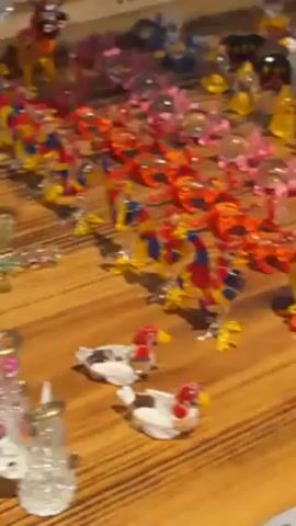 The video shows a variety of small trinkets displayed on a wooden surface. The trinkets are arranged in rows and consist of different colors and shapes. Some appear to be figurines, others resemble charms or keychains, while others are small bottles filled with glitter. The wooden surface has a natural grain and is likely a table or display case. The video quality is slightly blurry.