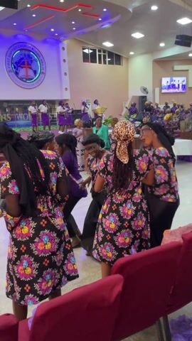 The video shows a group of women dancing in a church during a service or event. Several women are wearing dresses with a black background and vibrant floral patterns. Some have head coverings; one has a leopard print headscarf. They are smiling and interacting with each other as they dance. A choir or musical group is performing on a stage in the background, and their performance is also being shown on a screen. The church interior has burgundy chairs, and a sign or logo for "First Baptist Church Kubwa Abuja" is visible on the wall. The atmosphere appears lively and joyous.