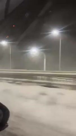 The video shows a view from inside a moving car during a snowstorm at night. Streetlights illuminate the snow-covered highway and nearby structures. The windshield wiper is visible, moving to clear the snow. The road surface and surrounding landscape are covered in white snow.