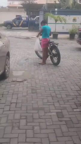 A person is standing next to a bicycle with wide tires. The person is wearing a blue t-shirt and pink shorts. They are holding a white plastic bag in their hands. The bicycle has a rack over the rear wheel. The person is facing away from the camera. In the background, there is a white van, a dark SUV, and a blue and yellow truck with people in it. There is also a fence and some potted plants. The ground is paved with gray bricks. The scene appears to be outdoors in a parking area or courtyard.