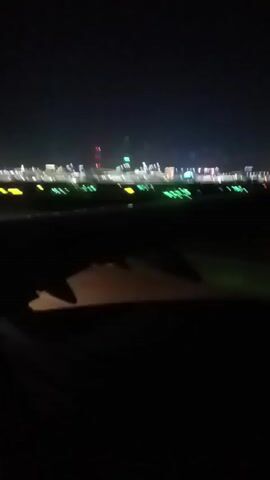 From inside an aircraft at night, we see blurred lights outside a window.  The wing of the plane is visible in the foreground.  The blurry background lights suggest an airport environment.  The scene is dark.