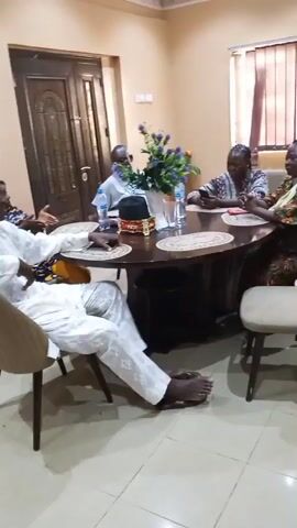 In a sparsely decorated room, four people are sitting around a dark, round table. The table has a bouquet of purple flowers in the center, as well as several water bottles. The person nearest the camera is a dark-skinned man wearing a white outfit; he is barefoot, leaning back in his chair with one arm on the table. Across from him, a dark-skinned woman in a brown and red outfit is sitting at the table. Two dark-skinned men sit to her left. The room has plain, light colored walls and a brown door can be seen in the background. Everyone is engaged in conversation.
