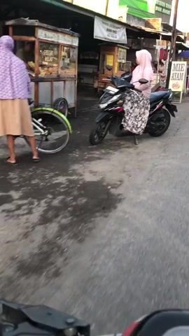 The media file captures a street scene in Balonggabus, Indonesia. In the foreground, a person wearing a purple patterned hooded jacket and a tan skirt is seen from the back, standing near a food stall. To their right, a woman in a pink hijab and a floral patterned dress is seated on a black motorcycle.

The food stall appears to be made of wood and glass, displaying various food items. A bicycle is parked next to the stall. Behind the motorcycle, a sign reads "MIE AYAM," which translates to "chicken noodles" in Indonesian, suggesting the type of food sold. The ground is paved and appears wet in some areas, possibly indicating recent rain or a cleaning.

The lighting suggests it is either early morning or late afternoon. The overall atmosphere seems to be that of a typical day in a marketplace or a street with local vendors. There are no strong indicators of specific emotions, but the scene conveys a sense of daily life and commerce. The background shows more stalls and buildings, hinting at a bustling local area. The presence of the motorcycle and the food stall points to a street food or small business setting.