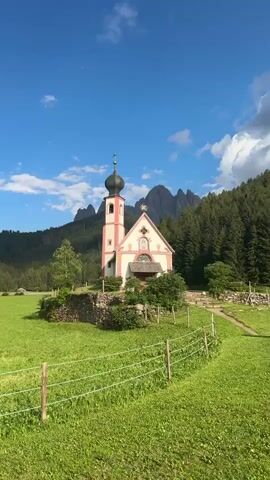 The video shows a small, white church with a pink roof and a dark steeple, set in a lush green field. The church has a single door and a few windows. Behind the church, a dense forest of evergreen trees covers a steep mountain slope. In the distance, jagged, dark mountain peaks pierce a bright blue sky dotted with fluffy white clouds. A simple wire fence runs across the foreground, separating the viewer from the field. The scene is serene and picturesque, bathed in sunlight.