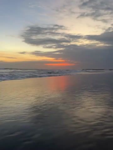 A low-angle shot of a beach at sunset. The ocean is calm with small waves gently lapping the shore. The sky is filled with dramatic clouds, with a bright orange glow from the setting sun peeking through. The wet sand reflects the colors of the sky, creating a beautiful, serene scene. The camera remains stationary throughout the video.