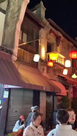 At night, a street is crowded with people. Above them, strings of red, yellow, and white lanterns are suspended, illuminating the street. The buildings on either side have aged, stone facades, and some have awnings in front of them. People are walking in both directions, forming small clusters as they talk or navigate the crowd. Some are wearing winter coats, while others are dressed more lightly. The lighting creates a warm, festive atmosphere on the street.