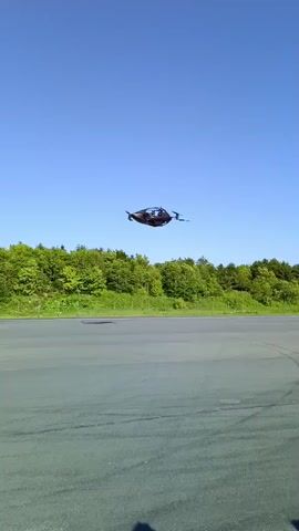 A black drone with four propellers is in flight over an empty asphalt surface under a clear blue sky. A line of green trees forms the background. The drone appears to be at a moderate altitude.