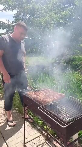 A man is barbecuing meat on skewers over a grill. He is wearing a dark t-shirt and shorts, with sandals on his feet. The grill is full of meat, and smoke is rising from it. The scene is set outdoors, with lush green foliage in the background. The sunlight suggests it is daytime, likely a sunny day given the brightness. The overall atmosphere appears relaxed, consistent with a casual outdoor meal. The context provided indicates the location is Vyazovka, Russia. There is no visible text in the media file.