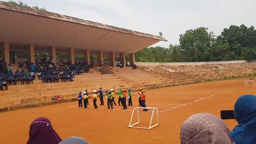 The video shows an outdoor sports stadium in Kalapet, India. The setting is a bright, overcast day, suggesting it is daytime. The stadium features tiered concrete seating filled with a large group of people, mostly young individuals dressed in blue. On the reddish-brown dirt field in front of the seating, a group of children, presumably students, are gathered. They are dressed in brightly colored sports attire and appear to be participating in a game or activity, holding what look like plastic bats and balls. Some are wearing helmets. The children are arranged in a line and seem to be listening to instructions from an adult who is partially visible. A white goal-like structure is set up on the field. Trees form a backdrop behind the stadium. The overall mood appears to be one of a school sporting event or a children's sports day. No visible text is present in the video.