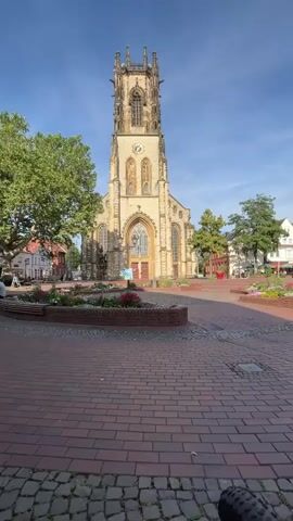 The video shows a wide shot of a European town square with a large, ornate church as the focal point. The church is made of light-colored stone and features a tall, Gothic-style tower with spires and arched windows. A clock is visible on the tower. In front of the church is a paved square made of red brick. There are several flower beds with colorful flowers and greenery, and some benches. Trees are scattered throughout the square. The sky is blue with a few wispy clouds. The lighting suggests it is daytime, possibly morning or late afternoon due to the long shadows. There are no people clearly visible in the foreground or the immediate vicinity of the church, though there may be some indistinct figures in the background near buildings on the edge of the square. The overall impression is a peaceful, picturesque urban scene.