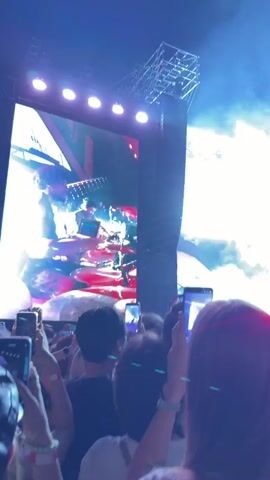 The video captures a live concert from the perspective of the audience. In the foreground, the backs of several audience members are visible, many holding up their smartphones to record or photograph the stage. The lighting is dynamic, shifting from purple and blue to bright red. On a large screen on the left, a drummer is shown playing. Above the stage, lighting rigs and speakers are visible. The word "PARADE" is lit up in white text on the right side of the stage. The overall atmosphere appears energetic and engaging.