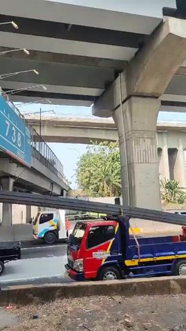 The video shows a series of trucks and cars driving under an elevated highway structure. The camera is positioned at ground level, looking upwards towards the highway. The highway is made of concrete beams and pillars. In the foreground, there is a truck with a blue and red cab and a long metal load. Further in the background, there is a white truck with a red container, a green truck with a blue tarp covering its load, and a yellow car. The environment appears to be urban, with buildings and trees visible in the distance. The lighting suggests it is daytime.