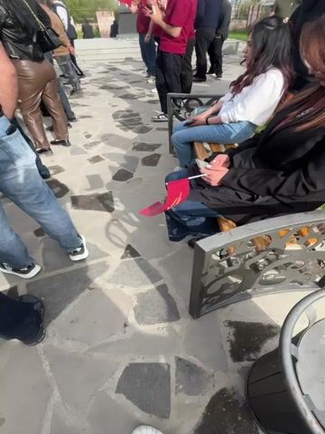 The video shows a group of people gathered in an outdoor area paved with irregular stone slabs. Several people are holding Armenian flags, and a few are sitting on benches. In the background, there is a silver statue of a woman holding a sun, and decorative stone pillars topped with eagle sculptures. Some people are standing, while others are walking around. The overall atmosphere seems to be one of a gathering or demonstration. The sky is overcast, indicating it might be a cloudy day.