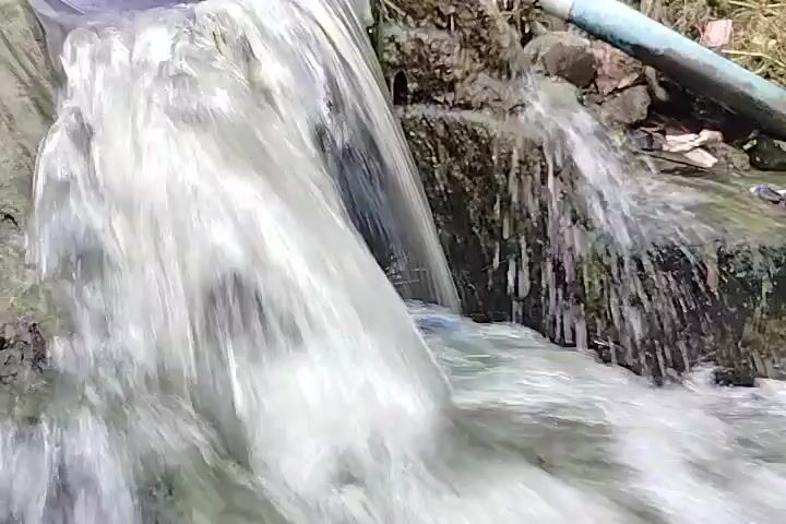 The video shows a close-up view of murky water flowing over rocks and debris. The water appears to be a mixture of clear and cloudy, with some white foam visible. The rocks are dark and wet, and there are bits of trash and plant matter scattered around them. A blue pipe is visible in the background, and it appears to be discharging water into the stream. The overall impression is one of a polluted or natural environment, with the focus on the movement of the water.