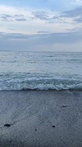 The video shows a calm sea lapping against a gray sandy beach under a cloudy sky. The small waves create white foam as they reach the shore. In the distance, there is a dark, possibly floating object on the water. The beach is speckled with pebbles and small debris.