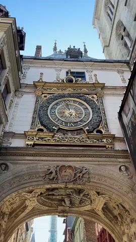 The video shows a low-angle, upward shot of a large, ornate clock on the facade of a building. The clock has Roman numerals and a sunburst design in the center. Below the clock is an arched entryway with detailed carvings. In the background, through the arch, a street with buildings and a tall, slender tower is visible. The sky is clear and blue. The video pans slightly up and then holds steady on the clock and archway.