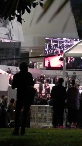 The video depicts a crowd of people at an outdoor event, likely a premiere or film festival. In the foreground, silhouettes of people stand on a patch of grass, looking towards a large screen that displays live footage of a red carpet event. There are also people visible on an elevated platform, watching the event. A white tent provides shelter nearby. The background features a building with large posters and billboards. The scene appears to be at night, and the lighting is artificial.
