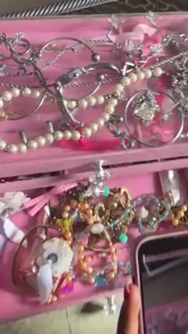 The video shows a two-tiered pink jewelry box overflowing with jewelry. The top tier contains silver-colored jewelry, including necklaces with pearls, chains, and pendants. The bottom tier is filled with gold-colored bracelets, necklaces with colorful beads, and decorative elements like fabric flowers and ribbons. A cell phone is visible in the lower right corner of the frame, apparently filming the jewelry box.