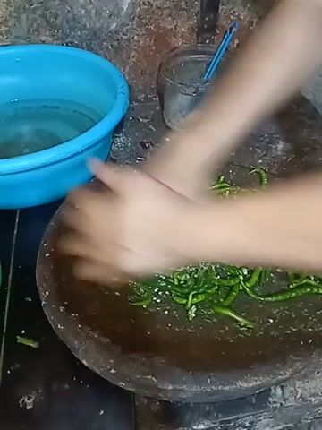 The video shows a close-up of a person's hands crushing green chilies in a stone mortar. The chilies are long and thin, and appear to be fresh. The person is using a pestle to mash the chilies, and there are some white specks, possibly salt, mixed in with the chilies. To the left of the mortar is a blue plastic bowl filled with water. To the right of the mortar, there is a small glass jar with a blue lid, containing what appears to be some chopped ingredients. The background is a rough, textured wall, possibly stone or concrete. The lighting is dim, suggesting an indoor setting, likely a kitchen. The action is focused on the preparation of the chilies.