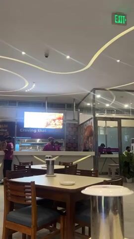 The video shows the interior of a restaurant. In the foreground, there are wooden tables and chairs. In the background, there is a counter with employees and a large screen displaying food. People are seen walking in and out of the restaurant, with one person in a blue shirt and another in a purple shirt standing near the entrance. The restaurant has modern lighting and decor, with a prominent "EXIT" sign on the ceiling.