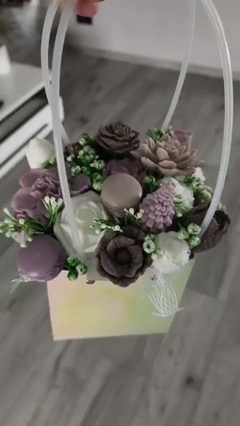 The video shows a person holding a small, iridescent gift bag filled with artificial flowers. The bag is square and has two white handles made of plastic tubing. The flowers are mostly in shades of purple and brown, with some white accents. There are also a few macarons and pinecone-shaped decorations interspersed among the flowers. The background is a wooden floor. The camera pans around the gift bag, showing it from different angles.