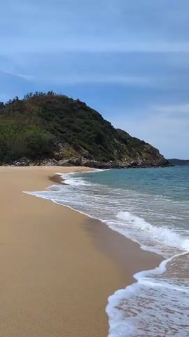 A beach scene with waves gently lapping against the shore. A lush green hill is visible in the background. The sky is blue with scattered clouds. The sand is a light golden color. The water is a clear turquoise.