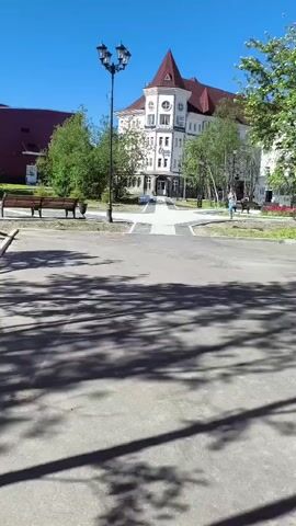 A person walks toward the camera on a paved path in a park. They are wearing blue jeans and a gray shirt. The path is bordered by green grass and trees. In the background, there is a multi-story white building with a red roof. The sky is clear and blue. A lamppost with three lights stands to the left of the path. Benches are visible on either side of the path. The ground in the foreground is paved and has shadows from trees.