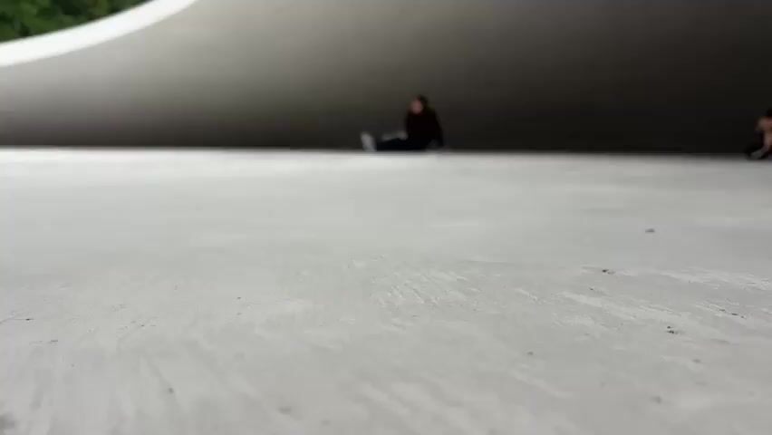 A person sits on a concrete floor in front of a large, curving concrete structure. The person is in the background and is out of focus, wearing a dark hoodie and jeans, with their legs extended. The camera is low to the ground, focusing on the texture of the concrete. Trees are visible in the upper left corner of the frame, suggesting an outdoor location. The lighting is overcast.