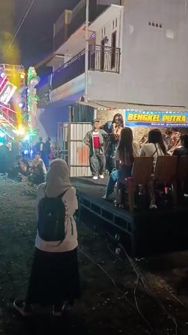 A video captures a nighttime outdoor performance. A young woman, wearing a hijab and a backpack, stands facing away from the camera, looking towards a raised stage. The stage has a black platform and is occupied by two performers. One, a person wearing a hat and a gray jacket over a red shirt, stands behind a microphone stand. The other, a woman with long dark hair, is also holding a microphone and appears to be singing. Several other individuals are seated behind the performers, some watching the performance. To the left of the stage, a large sound system is set up with colorful stage lighting, casting beams of light into the air. In the background, a building with white walls and balconies is visible, and a sign reads "BENGKEL PUTRA RIA." The ground appears to be dirt or gravel, and cables are laid out across it. The overall scene suggests a lively outdoor event or concert.