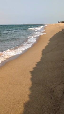 The video depicts a tranquil beach scene. Golden sand stretches along the shore, meeting the edge of a blue-green ocean. Gentle waves with white foam crest and recede along the shoreline. In the distance, a few people can be seen scattered along the beach, and some appear to be in the water. The sky above is a pale blue, suggesting a clear day. A distinct shadow line divides the foreground sand, adding depth to the composition.