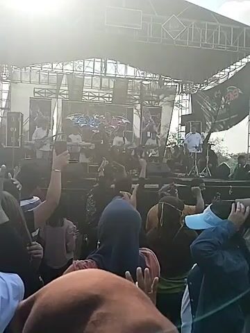 A crowd of people is enjoying a live music performance outdoors. The band is on a large stage, complete with a backdrop featuring their name. Several crowd members are dancing and filming the performance on their phones. The environment suggests a sunny day, potentially a public event or festival.