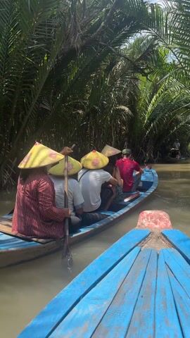 The video shows a point-of-view shot from a blue boat following another blue boat on a narrow river through a tropical forest. In the boat ahead, multiple people sit and paddle through the murky water. The people wear conical hats, and a woman in a red and white striped shirt is actively rowing. The dense green vegetation, including palm trees, lines both sides of the river, forming a lush canopy. The sunlight filters through the leaves, creating patches of light and shadow on the water and surrounding foliage.