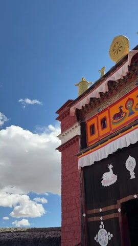 The video captures an upward view of a traditional Tibetan Buddhist temple building against a bright blue sky with scattered white clouds. The temple features red brick or stone walls and an elaborately decorated roof. Golden ornaments, including a Dharma wheel and what appear to be two deer or gazelles flanking another golden figure, adorn the rooftop. Below the roofline is a colorful frieze with intricate designs in orange, red, and yellow hues. Dark and white fabric drapes, decorated with white symbols such as deer/gazelles and other Buddhist motifs, hang from the building's facade. A low stone wall is visible in the bottom-left foreground. The scene is bright and sunny, highlighting the intricate details of the architecture. There are no people visible, and the only movement is the subtle sway of the fabric drapes in the breeze.