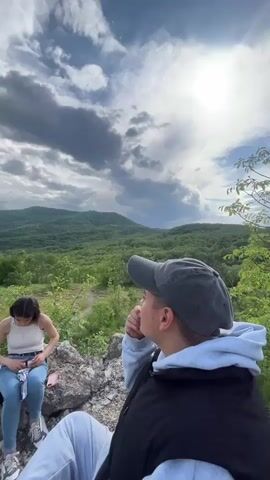 A medium shot shows a sunny day with a view of a forested hill or small mountain range. In the foreground, a woman in jeans and a white tank top sits on some rocks looking down at something in her hands. To the right of her, a person in a grey baseball cap and grey hoodie looks off towards the hills. The clouds are moving quickly in the sky.