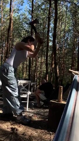 A woman in a white tank top and baggy gray pants is chopping wood with an axe in a forest clearing. She is positioned to the left of a large log and swings the axe with force, creating a spray of woodchips. A second person, wearing a dark shirt and shorts, is partially visible sitting on a chair behind the woman and the log. Several other chairs and a hammock are present in the background, along with tall pine trees and dappled sunlight.
