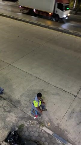This media file shows a high-angle shot of a person squatting on a small, blue stool on a paved area. The person is wearing a blue shirt, jeans, and a bright yellow safety vest over a grey vest. They appear to be male and have dark hair. The ground is concrete with visible cracks and lines. To the left of the person, there is a motorcycle parked and some debris. In the background, a white truck is blurred due to motion, suggesting it is moving on a road. The lighting suggests it is nighttime or dusk, with artificial lights illuminating the scene. The overall atmosphere seems ordinary, possibly a roadside or a work area. There are no visible text elements in the frame. The setting appears to be an urban or semi-urban environment, possibly in Pekalongan, Indonesia, given the context. The person's posture suggests they might be resting, waiting, or observing something.
