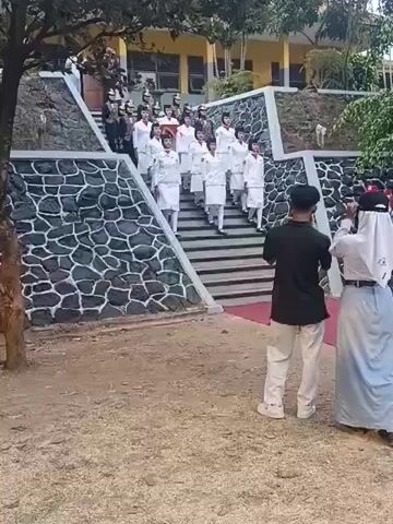 A group of young people in white uniforms with red accents, some wearing hats, are descending a set of stairs. The stairs are flanked by stone walls with a decorative pattern. At the bottom of the stairs, two individuals are facing away from the camera, one holding a phone up as if taking a picture. Behind the stairs, a yellow building with windows is visible, suggesting a school or similar institution. The ground in the foreground is a mix of dirt and gravel. The scene appears to be outdoors during the daytime.