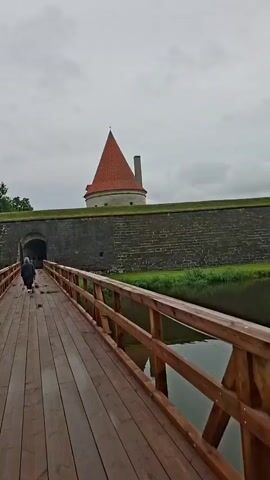 A person walks across a wooden bridge towards an arched entrance in a stone fortress wall. The fortress has a conical red-roofed tower and is surrounded by a moat. On the other side of the moat, a large, traditional European-style house with a red roof stands amidst lush greenery. The sky is overcast, suggesting a cloudy or potentially rainy day. The overall mood is peaceful and historical, with the fortress and the old house evoking a sense of a bygone era. The scene is set in a location with historical architecture, likely a tourist attraction or a preserved historical site. No text is visible in the media file.