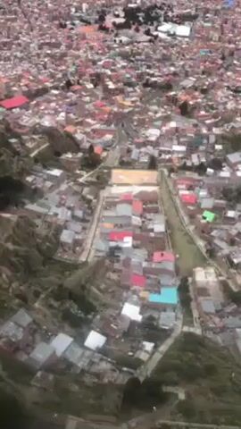 The video displays an aerial view of a densely populated urban landscape. The scene is dominated by closely packed buildings with a mix of roof colors, including red, blue, and grey. A central open space, resembling a long park or avenue, cuts through the dense architecture. The terrain is somewhat hilly, with greenery visible on the slopes. The overall impression is one of a crowded, sprawling city viewed from a high altitude.