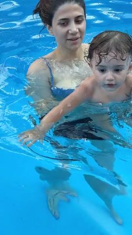 The video shows a woman holding a young child in a swimming pool. The woman is wearing a blue swimsuit and has her hair pulled back. The child is naked and has curly brown hair. Both are submerged in clear blue water. The woman appears to be talking to the child, who is looking at her with a curious expression. The lighting suggests it is daytime. The scene takes place in a swimming pool, likely an outdoor one given the bright lighting and visible reflections on the water's surface. There are no visible objects other than the people and the pool water. The video is short and appears to capture a moment of interaction between the woman and child in the pool. There is no visible text in the video.