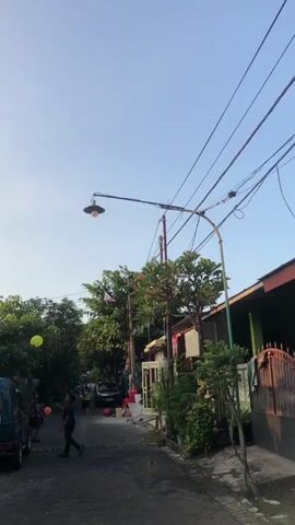 A street scene with a clear blue sky and scattered clouds. A streetlamp extends over a narrow, paved street lined with houses and lush trees on both sides. A few cars and a motorcycle are parked along the street. Two individuals, one holding a basketball and the other a balloon, are visible in the distance. The scene appears to be a residential area, possibly a neighborhood or alleyway, with a generally calm and quiet atmosphere.