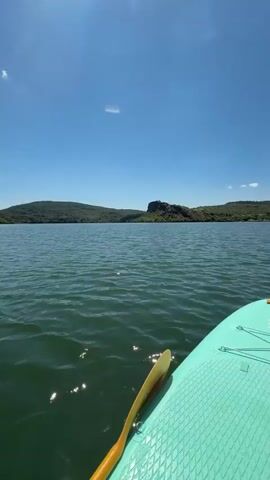 A first-person view from a stand-up paddleboard shows a paddle resting on the board as it moves across a green, rippling body of water under a bright blue sky with a few wispy clouds. The paddleboard is a light teal color with orange bungees and a yellow paddle handle visible. In the distance, a tree-covered shoreline stretches across the horizon, with a prominent rocky outcropping visible on the left side of the frame in the initial moments of the video. The sun creates lens flares in the upper left corner of the frame. The camera is pointed forward, capturing the expanse of the water and the distant landscape.