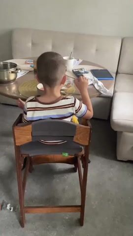 A young boy with short brown hair sits in a wooden high chair facing away from the camera. He is wearing a striped t-shirt with blue, white, brown, and black horizontal stripes. The boy is at a round table, eating with a utensil. On the table are a pot, papers, a cup, a remote control, and a tablet. Behind the table is a beige sectional sofa. The floor appears to be concrete.
